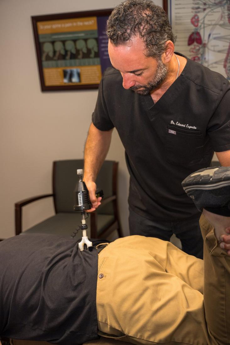 Dr. Edward Esposito, whose name is embroidered on his scrubs, applying a handheld adjusting instrument to a patient's back.