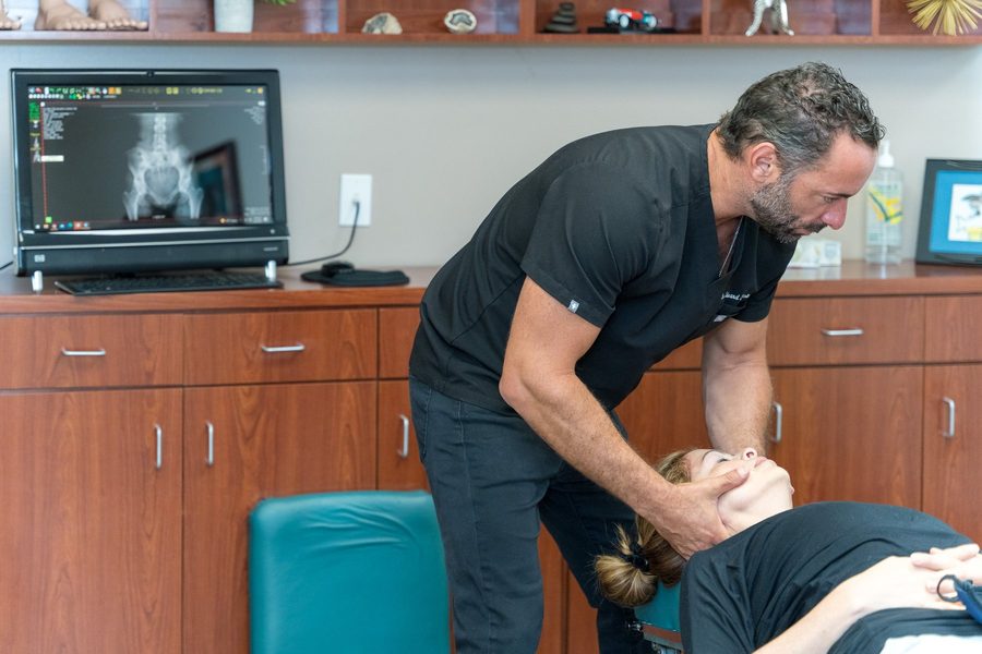 Dr. Esposito performing a cervical adjustment with a patient's imaging displayed on screen behind him.